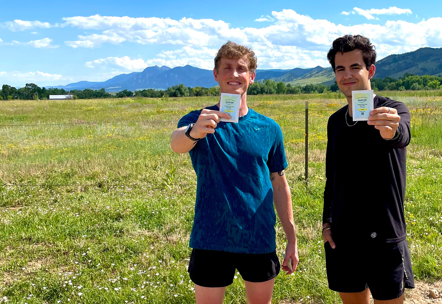 Two male runners holding up sample packs of Enerza performance drink mix for endurance athletes. Grassy open space with mountains in the background.